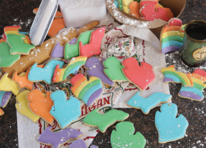 A messy kitchen with state of Michigan cut out cookies in various shades of the rainbow. Flour is scattered over the cookies, coffee mug, towel, rolling pin, and cardboard cookie carrier. The flour container lid is on its side, leaning against the flour container.